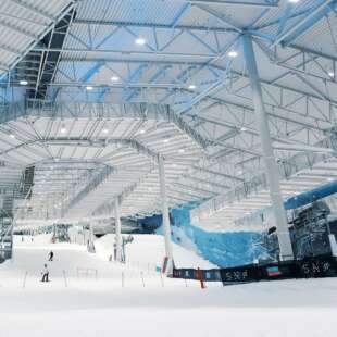 Intérieur industriel d'une station de ski avec piste enneigée, skieurs, passerelles en métal et mur bleu en arrière-plan.